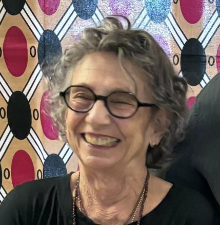 Lois Engelman, smiling, with silver curls and round dark glasses, in front of a patterned cloth