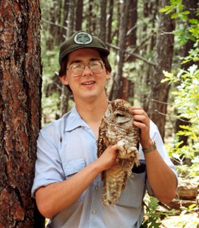 A young Jeff Jenness in a Forest Service cap, standing against a ponderosa pine, holding a Mexican spotted owl in both hands