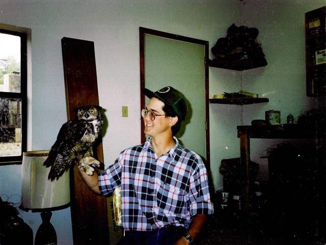 A young Jeff Jenness in a plaid shirt and cap, smiling at a great horned owl perched on his hand