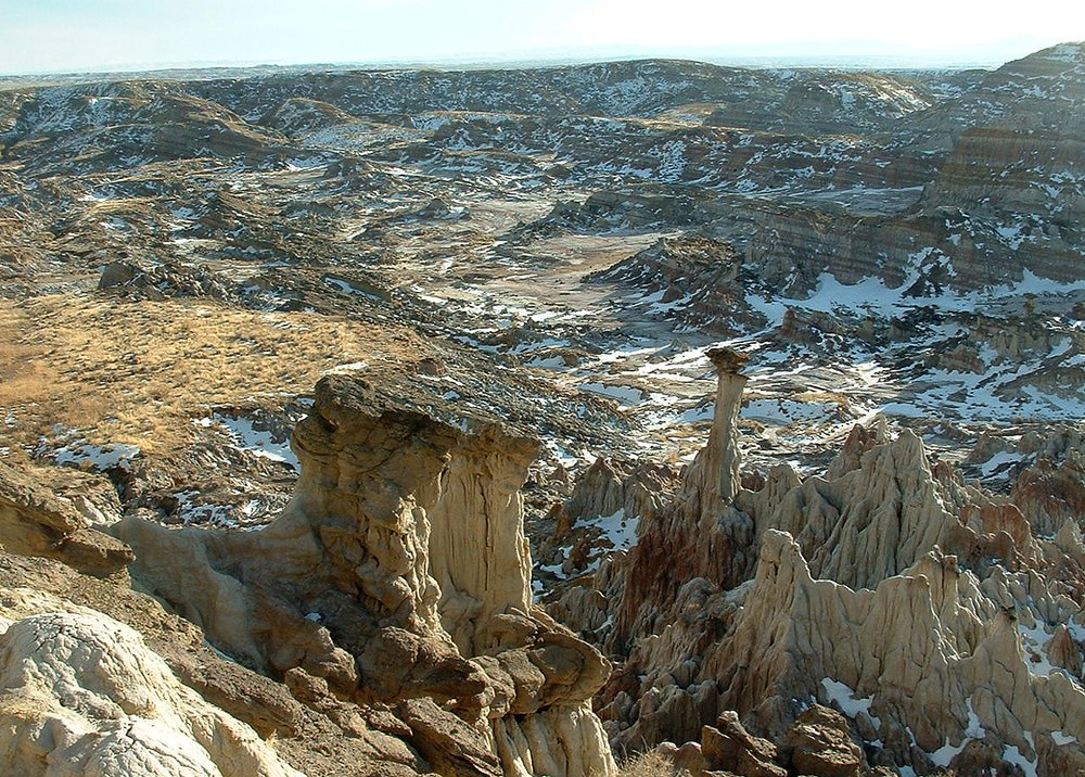 Hell's Half Acre, Wyoming: a wide badlands basin of eroded gray, tan and rust-colored rock, with a flat-topped hoodoo and a thin rock spire in the foreground, patches of snow in the hollows, and a prairie horizon under a pale sky